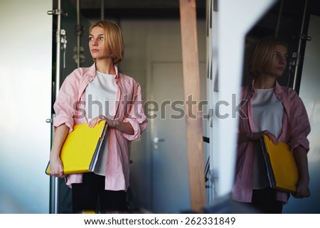 Portrait of young stylish woman holding bright yellow book standing near shelf in home interior, creative female designer with big magazine catalog standing in her studio while focused looking away