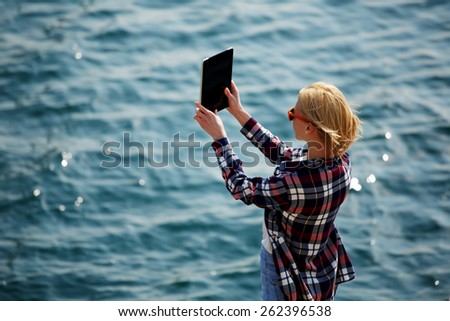 Back view of a woman taking picture with a digital tablet camera standing against sea background on the beach, female tourist standing on coastline photographing nature view, wing in sunny day