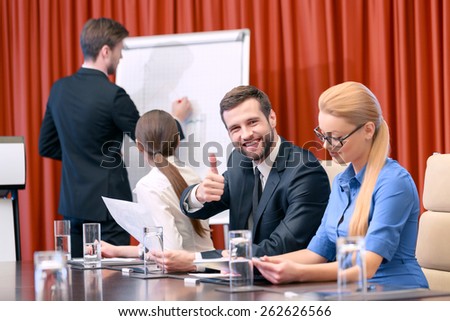 Great job. Businessman in classical black suit paints a chart on the flipchart while his female colleagues turned around to see his picture and his male partner smiles and shows his thumb up