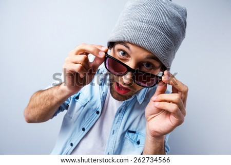 Hi there! Top view of funny young man adjusting his sunglasses and looking at camera while standing against grey background