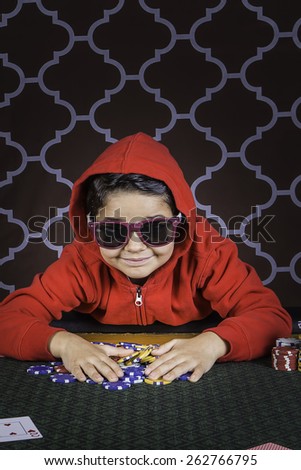 A young boy sitting at a poker table with a hoodie and sunglasses gambling playing cards with a brown background