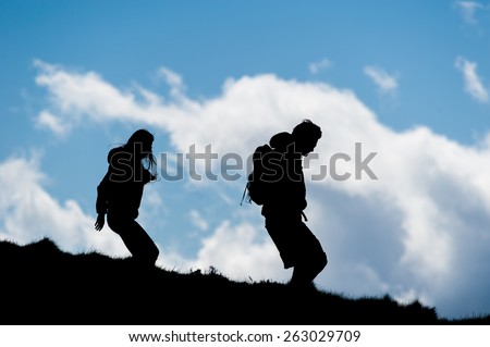 Silouette of a young man and woman walking down a hill against a blue sky with clouds.