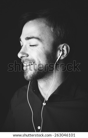 Close up head and shoulders greyscale portrait of a handsome cute charismatic guy listening to music on a set of earplugs with a smile of bliss and pleasure