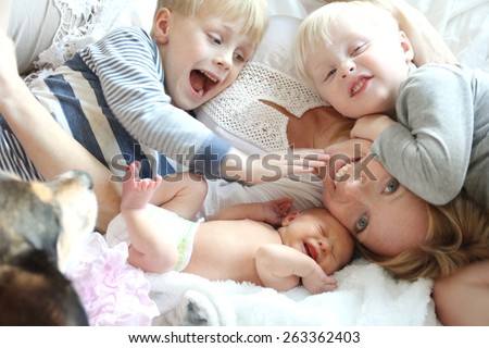 A happy young mother is laying on the bed in chaos, with her three young children, two wild boys and a newborn baby girl, as their pet dog sits by. Royalty-Free Stock Photo #263362403