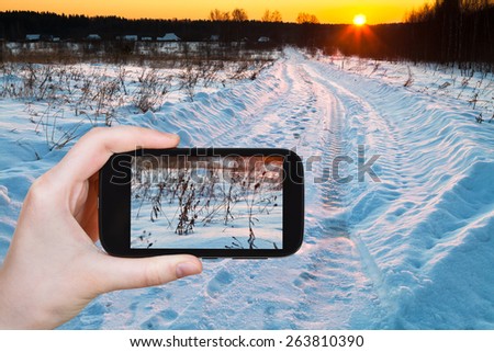 travel concept - tourist takes picture of sunset over blue winter snowdrifts on country field on smartphone,