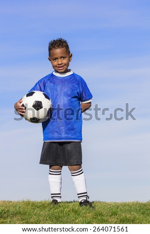 Young african american boy soccer player holding a ball standing on a grass field with a simple blue sky background. 