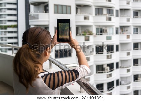 Woman holding tablet in her hands showing trees in the forest on the screen and builing in the background.