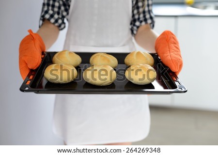 woman holding hot roasting pan with hot, freshly baked bread rolls. Protecs hers hands with orange kitchen gloves