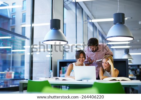 Group of international university students having fun studying in library, three colleagues of modern work co-working space talking and smiling while sitting at the desk table with laptop computer Royalty-Free Stock Photo #264406220