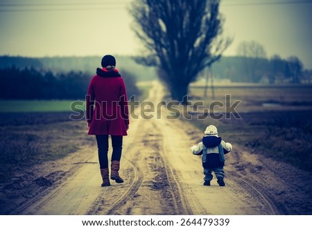 Mother with child walking by rural sandy road. Countryside under cloudy sky, photo with vintage mood. 