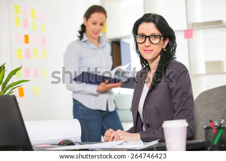 Businesswoman sitting at her desk with colleague on background