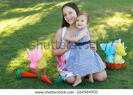 Two smiling girls sisters: baby and teen with wicker basket with chocolate eggs for Easter holiday on green grass in park
