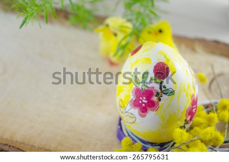 Hand painted decoupage Easter egg on a stand with yellow flowers and chickens in the background on a wooden table