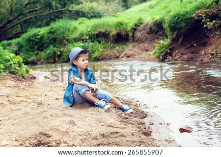 boy sitting on the sandy shore of the river, looks into the distance, holding a stone in his hand, wearing a blue jacket and hat