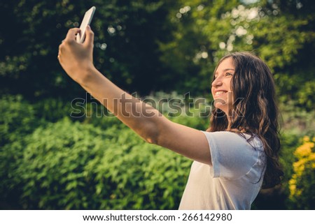 Pretty young caucasian woman smiling cheerfully for a selfie on her smartphone. Green natural environment in background. Filtered effects.