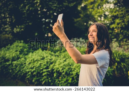 Pretty young caucasian woman smiling cheerfully for a selfie on her smartphone. Green natural environment in background. Filtered effects.