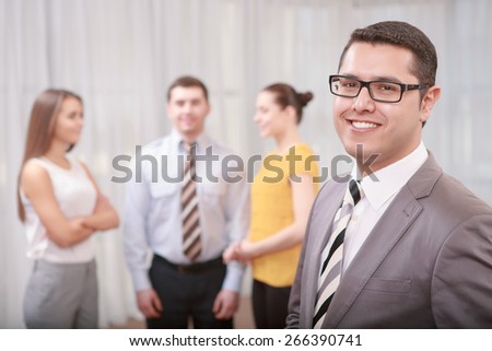 Team work. Young confident businessman in formalwear and glasses looking with a smile straight at a camera with his team discussing something on the background in blurry