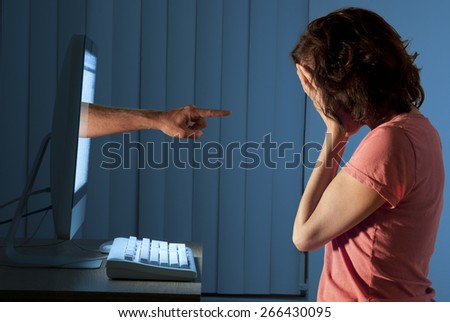 Severely distraught young woman sitting in front of a computer with a judgmental hand pointing at her from within the computer monitor cyber bullying her and social media stalking her. Royalty-Free Stock Photo #266430095