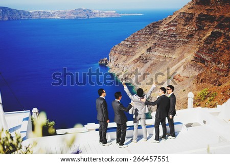 stylish happy rich asian groom with s?hafer and best men watching the blue sea on the island of Santorini before wedding ceremony