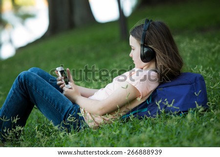 Girl lying on green grass outdoors in summer or spring reading her cellphone or tablet and wearing headphones