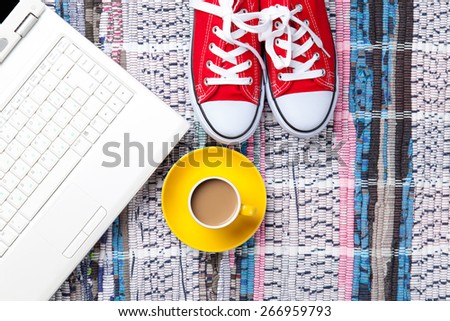 Cup of cappuccino near red gumshoes and computer on carpet background. Above view