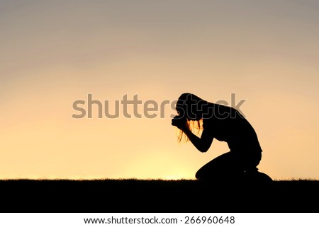 A silhouette of a young Christian woman is bowing her head in prayer, and desperation outside during sunset. Royalty-Free Stock Photo #266960648