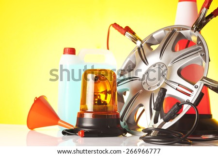 Car accessories - an alloy wheel with jump start cables, traffic cone, amber beacon light with car liquids and orange funnel on yellow background