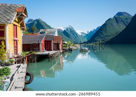 View over the fjord Fjaerlandsfjord and the village of Mundal (or Fjaerland) with some snow-capped mountains, a boat, reflection in the water and yellow and red boathouses in Sogn og Fjordane, Norway. Royalty-Free Stock Photo #266989451
