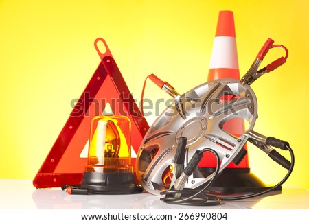 Car accessories - emergency road triangle with amber beacon light and jump start cables on an alloy wheel on yellow background