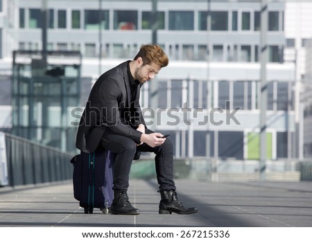 Portrait of a young man looking at mobile phone. Male traveler sitting on suitcase reading text message