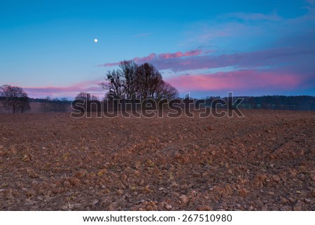 After sunset landscape of sky with moon and red clouds over plowed field.