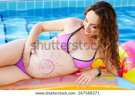 Beautiful pregnant woman with picture on her belly relaxed near swimming pool with blue water