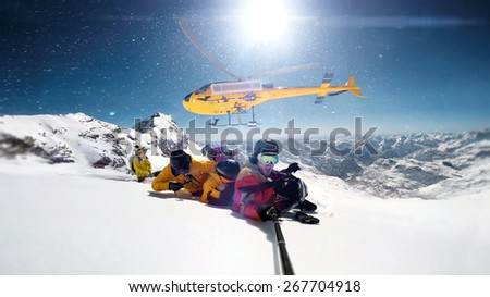 snowboarders were dropped by a helicopter at the top of the mountains while one person is taking a smile selfie with a wide angle camera.  The sun is shining brightly in the blue sky. 