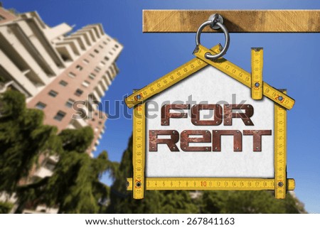 House For Rent Sign - Wooden Meter. Yellow wooden meter ruler in the shape of house with text for rent. For rent real estate sign on blue sky with tall and blurred buildings in the background