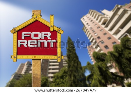 House For Rent Sign - Wooden Meter. Yellow wooden meter ruler in the shape of house with text for rent. For rent real estate sign with tall and blurred buildings in the background