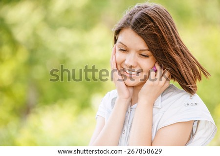 Portrait of young smiling brunette woman wearing white shirt and speaking on telephone at summer green park.