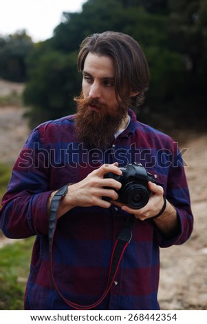 Young bearded man taking photo with his digital camera, handsome hipster tourist checking out the sights in nature landscape, hiker looking out across the nature holding his professional camera