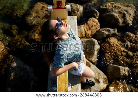 Top view portrait of cool young woman posing with her skateboard in front of the sea while enjoying summer sunshine, attractive hipster girl in sunglasses relaxing outdoors standing on rocks seashore