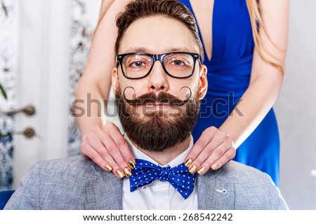 Woman with manicure straightens a bow tie to old-fashioned man in glasses with a beard and curled mustache. Photo toned in sepia, stylized retro shoot. Royalty-Free Stock Photo #268542242