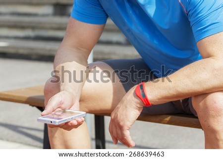 Sportive man sits on a bench and checks his fitness results on a smart phone. He wears a fitness tracker wristband on his left arm. Royalty-Free Stock Photo #268639463