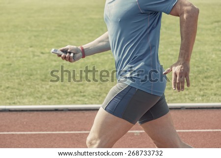 Runner in a stadium checks his fitness results on a smartphone while running. He wears a fitness tracker wristband on the right arm. Photo is filtered with slight antique effect.