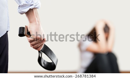 Family violence and aggression concept - furious angry man raised punishment hand holding leather belt over scared or terrified woman sitting at wall corner Royalty-Free Stock Photo #268781384
