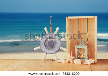old nautical wood wheel, shells and lantern on wooden table over sea background.
