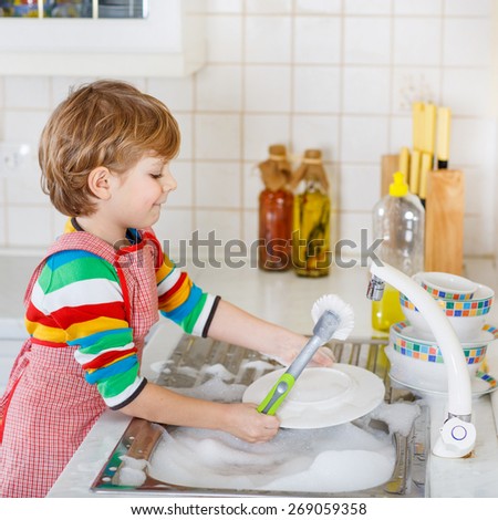 Adorable happy little blond kid boy washing dishes in domestic kitchen. Child having fun with helping his parents with housework. Indoors, kid in colorful clothes.