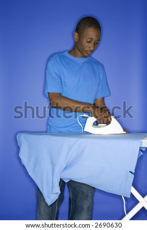 Portrait of African-American  teen boy ironing shirt standing against blue background.