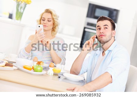 A picture of a young couple eating breakfast in the kitchen and using smart phone