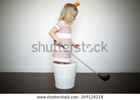 Adorable little girl playing in her room. Staying in the wicker basket. Fake golf club in her arms. Crown on her head.