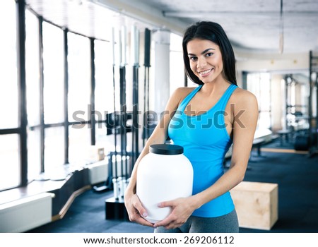 Happy woman holding plastic container with sports nutrition at gym and looking at camera