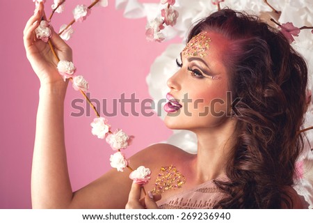 Fashion Art Beauty Portrait. Beautiful Girl with fantasy creative make up in spring garden holding flowering sakura branch. Pink background. Spring time concept