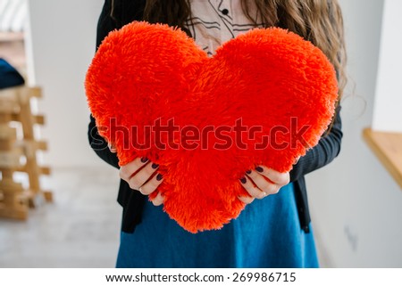 Picture of a big red heart in hands, female holds handmade sewn soft toy, woman with Valentine gift against the window, happy girl smiling, conceptual image of health care or love.